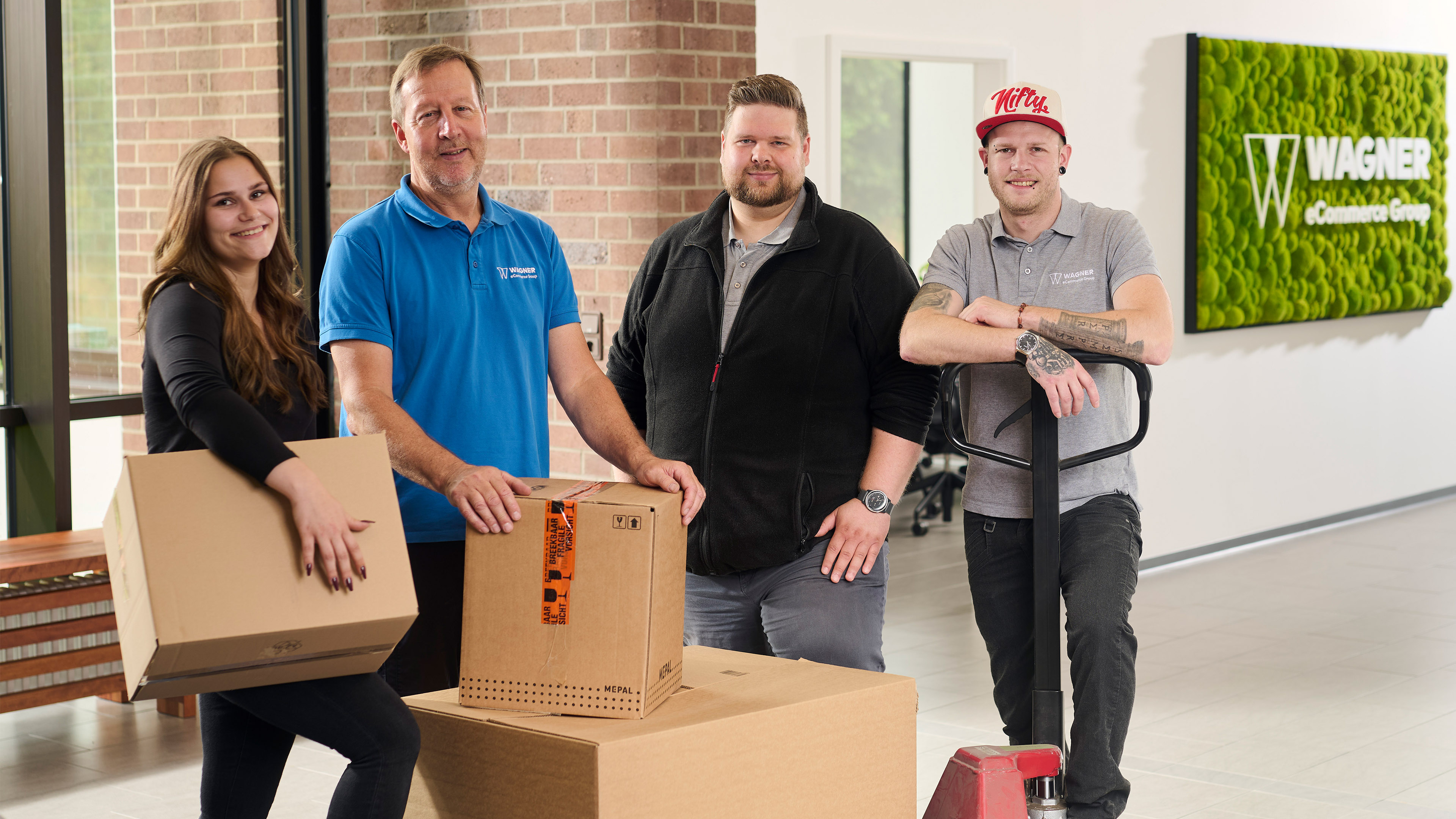 Das Bild zeigt vier Personen, die in einem modernen Bürogebäude vor einer Wand mit einem grünen Schild stehen, auf dem 'Wagner eCommerce Group' zu lesen ist. Links steht eine junge Frau mit langen braunen Haaren, die ein schwarzes Oberteil trägt und einen Karton in den Händen hält. Neben ihr steht ein älterer Mann in einem blauen Polo-Shirt mit dem Wagner-Logo, der ebenfalls einen Karton auf einem Tisch vor sich hält. Rechts von ihm steht ein Mann mit kurzem Haar und Bart, der eine schwarze Jacke trägt. Ganz rechts steht ein Mann in einem grauen Polo-Shirt mit dem Wagner-Logo und einer roten Baseballkappe, der einen Palettenhubwagen hält. Alle Personen lächeln in die Kamera.