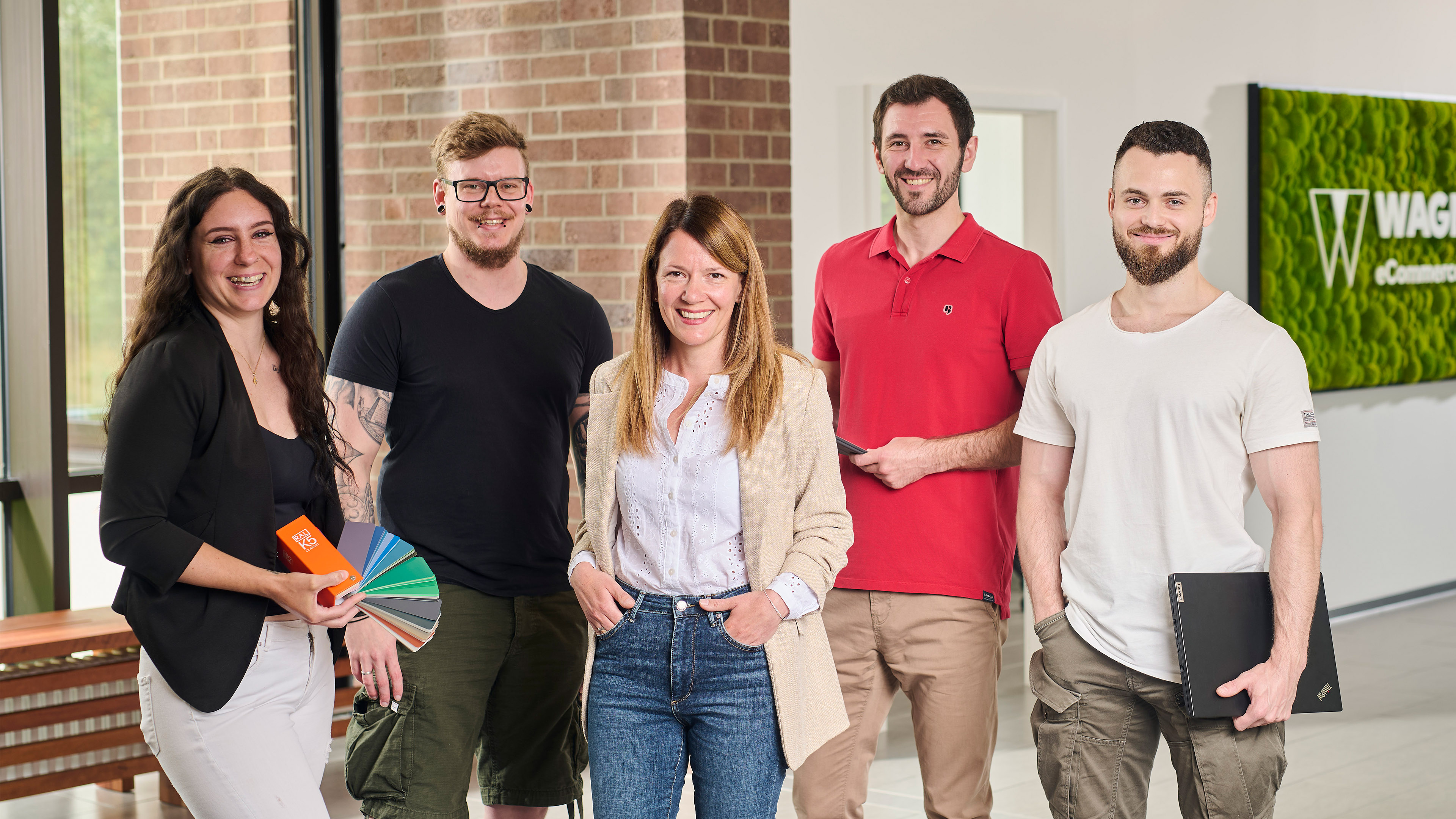 Das Bild zeigt fünf Personen, die in einem modernen Bürogebäude vor einer Wand mit einem grünen Schild stehen, auf dem 'Wagner eCommerce Group' zu lesen ist. Links steht eine Frau mit lockigem braunem Haar, die ein schwarzes Oberteil und eine weiße Hose trägt. Sie hält Farbpaletten in der Hand. Neben ihr steht ein Mann mit Brille und kurzem Haar, der ein schwarzes T-Shirt und olivgrüne Shorts trägt. In der Mitte steht eine Frau mit langen braunen Haaren, die ein beiges Sakko, ein weißes Hemd und eine blaue Jeans trägt. Rechts von ihr steht ein Mann mit einem roten Poloshirt, der ein Tablet in der Hand hält. Ganz rechts steht ein Mann mit kurzem Haar und Bart, der ein weißes T-Shirt und khakifarbene Hosen trägt und einen Laptop in der Hand hält. Alle Personen lächeln in die Kamera