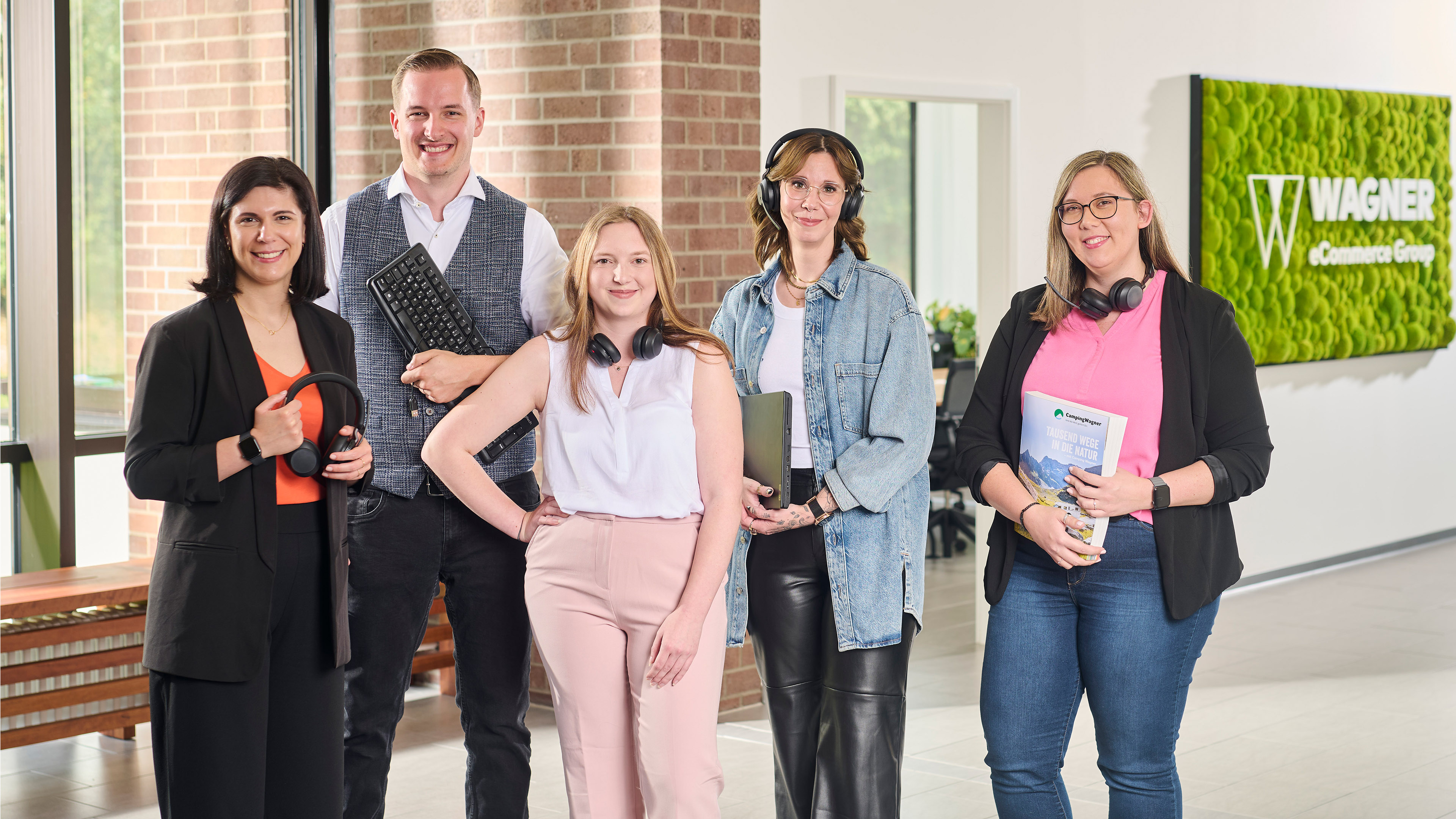 Fünf lächelnde Personen in einem Büro, halten Kopfhörer, Tastatur, Laptop und Magazin, vor einem Wagner eCommerce Schild.