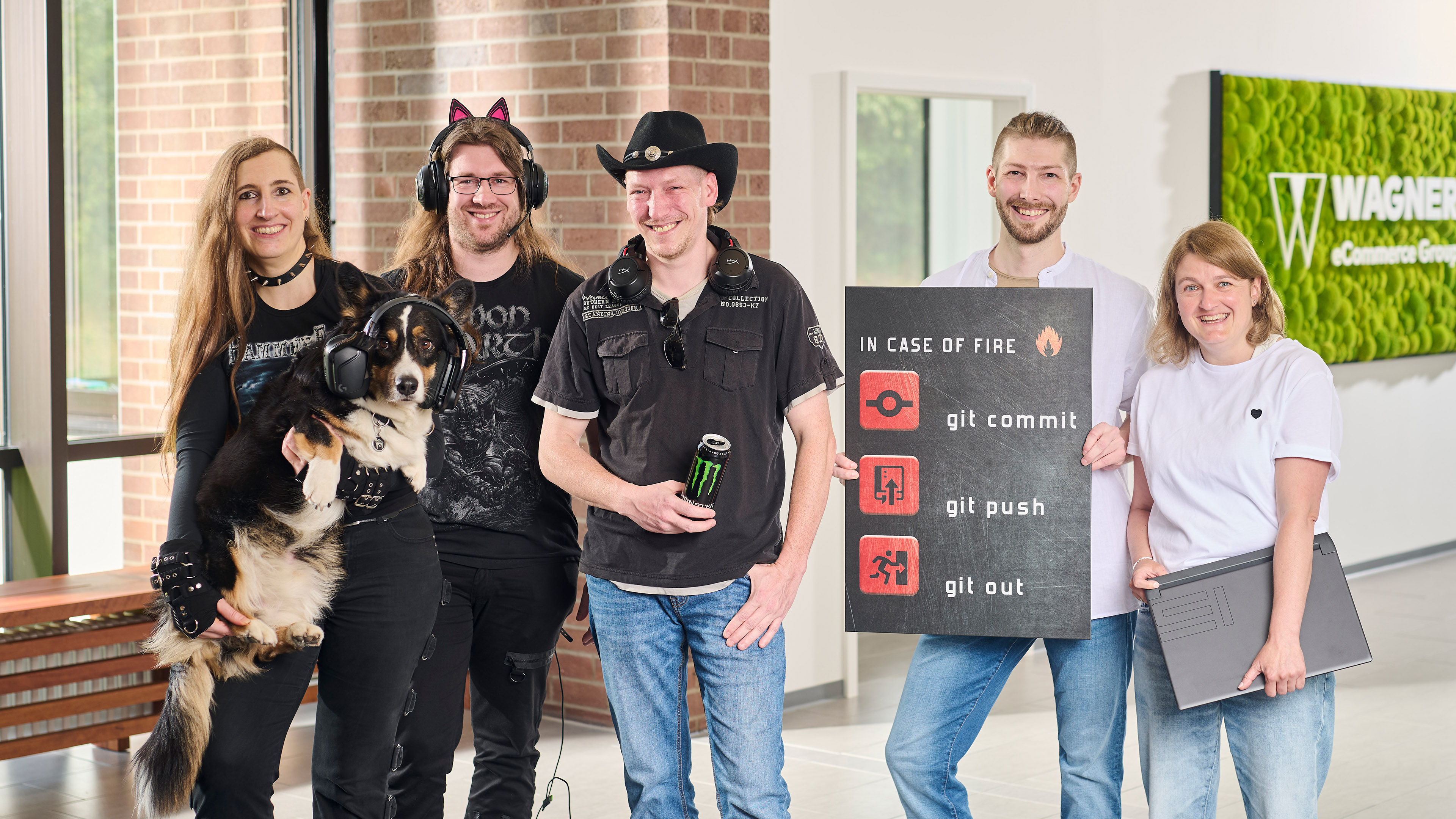 Das Bild zeigt sechs Personen, die in einem modernen Bürogebäude vor einer Wand mit einem grünen Schild stehen, auf dem 'Wagner eCommerce Group' zu lesen ist. Links steht eine Frau mit langen braunen Haaren, die ein schwarzes Metal-T-Shirt trägt und einen Hund hält, der Kopfhörer trägt. Neben ihr steht ein Mann mit langen Haaren und einer Katzenohren-Kopfbedeckung, der ebenfalls ein schwarzes T-Shirt trägt. Daneben steht ein Mann mit Cowboyhut und Kopfhörern um den Hals, der eine Dose Monster-Energy in der Hand hält. Rechts von ihm steht ein Mann, der ein Schild mit der Aufschrift 'In Case of Fire: git commit, git push, git out' hält. Ganz rechts steht eine Frau mit blonden Haaren und weißem T-Shirt, die ein Laptop unter dem Arm hält. Alle Personen lächeln in die Kamera.