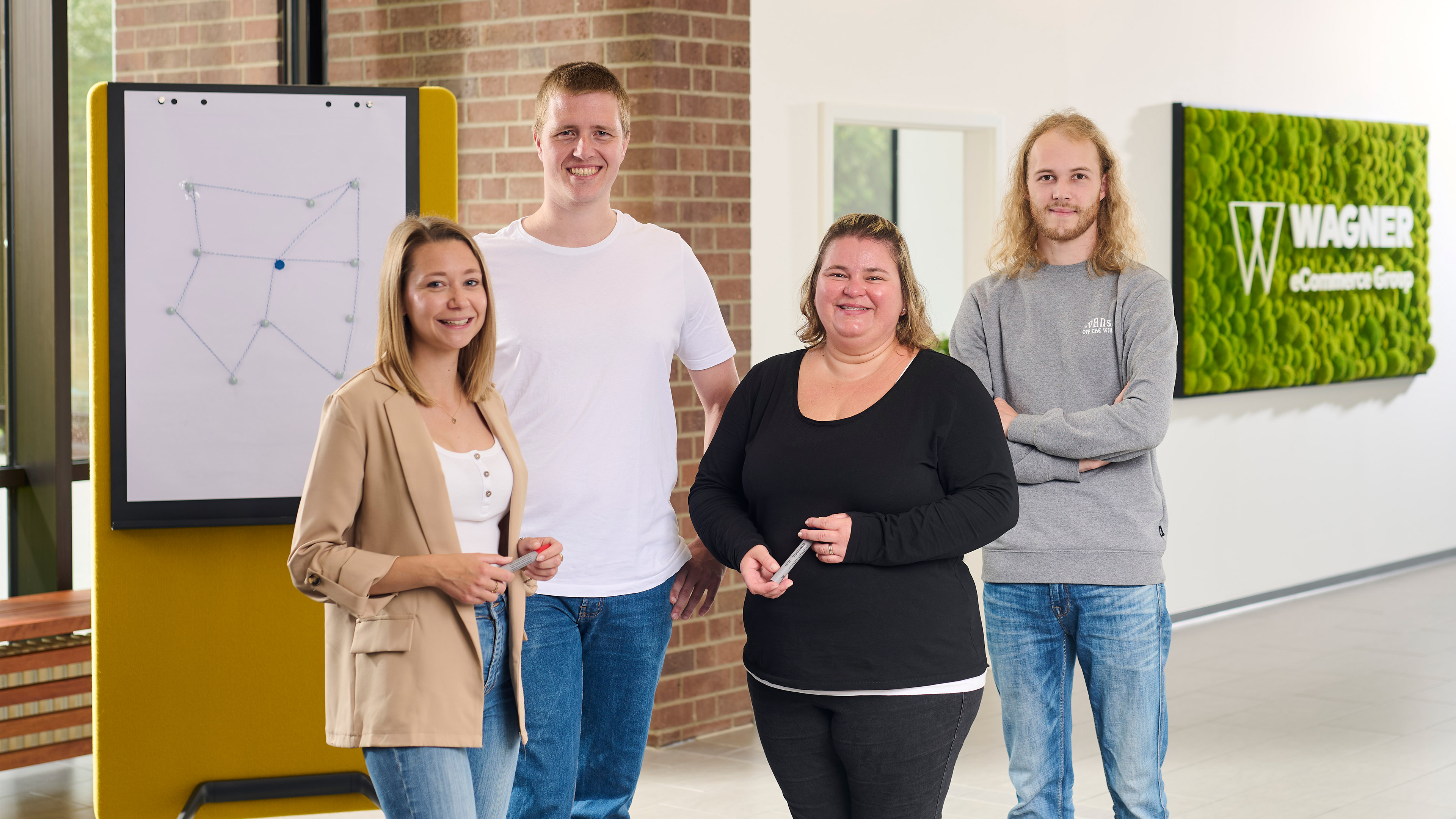 Eine Gruppe von vier lächelnden Personen steht in einem modernen Büro. Sie befinden sich vor einem Whiteboard mit einer einfachen geometrischen Zeichnung und einem grünen Wandschild, auf dem 'Wagner eCommerce Group' steht.