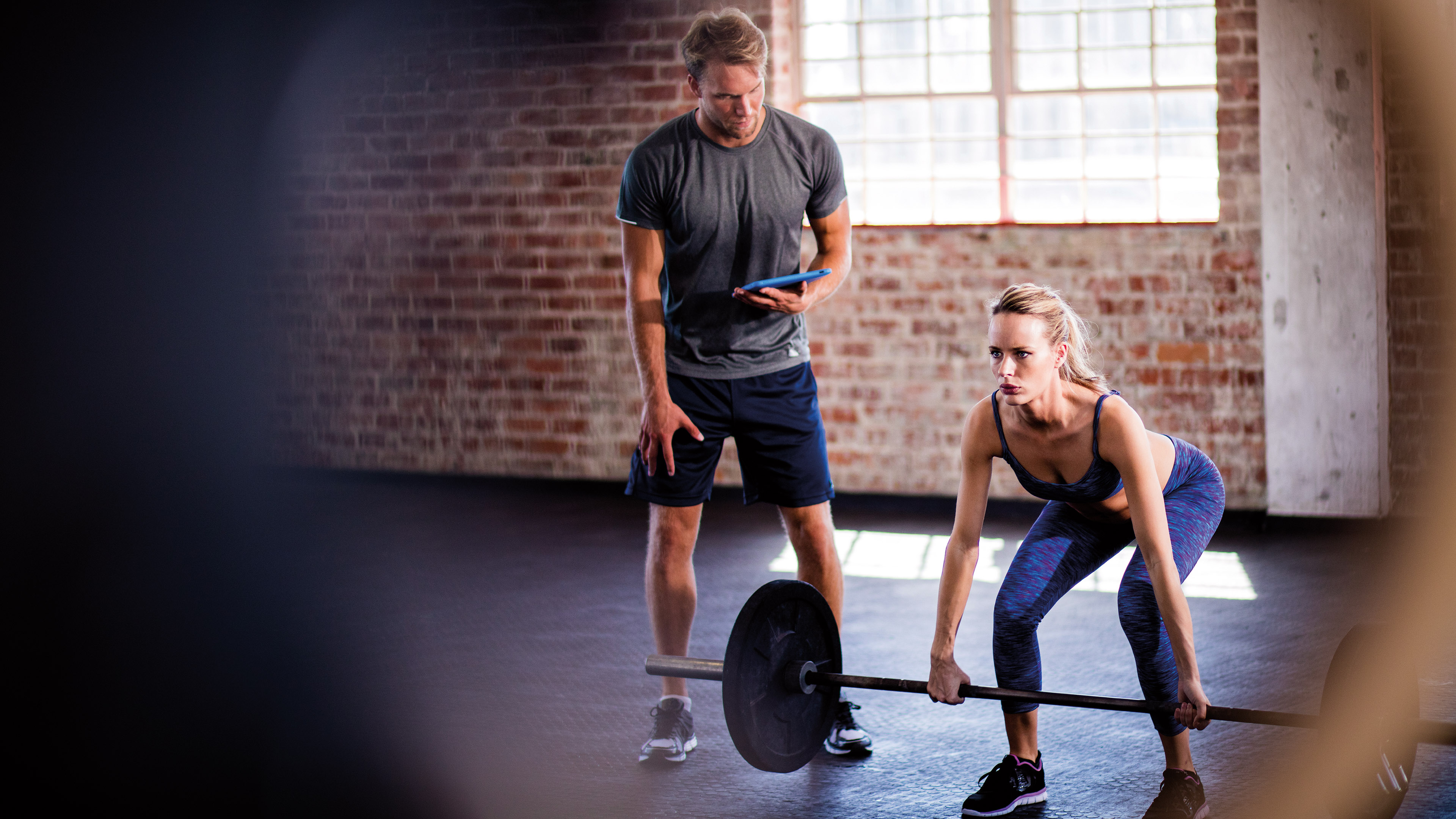 Trainer beobachtet eine Frau beim Gewichtheben in einem Fitnessstudio mit Backsteinwänden, beide konzentriert bei der Arbeit.