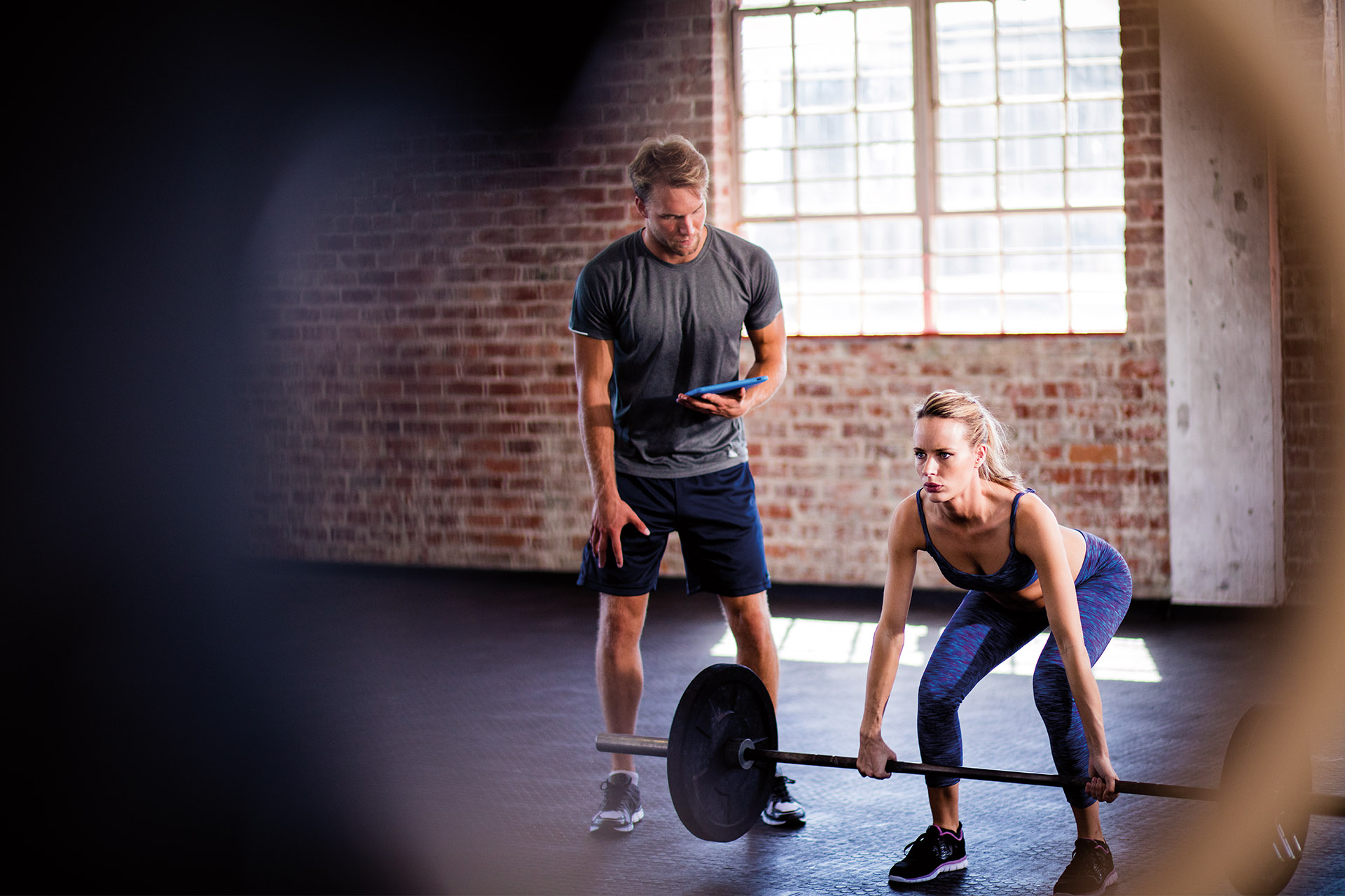 Trainer beobachtet eine Frau beim Gewichtheben in einem Fitnessstudio mit Backsteinwänden, beide konzentriert bei der Arbeit.
